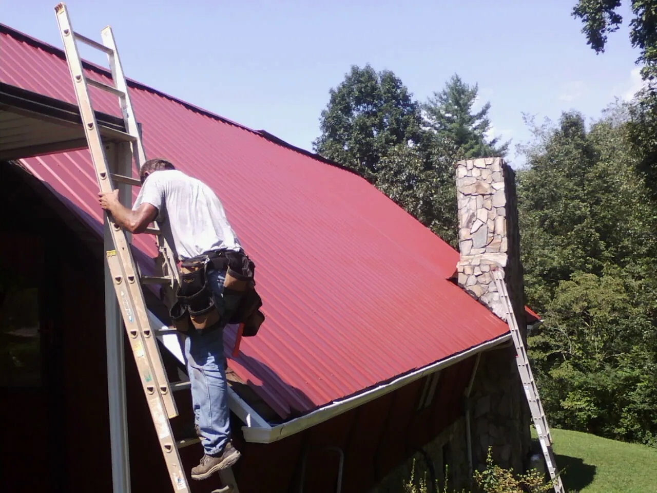 Roofer on a ladder beside a red metal roof and stone chimney — Soffit & Fascia Repair by Roofing Brothers in Livermore
